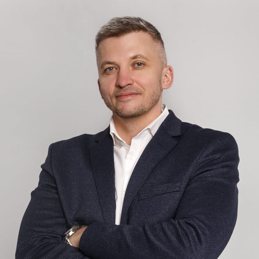 Professional-looking man with short, light hair, wearing a navy blue blazer and white dress shirt, smiling with arms crossed against a neutral gray background.
