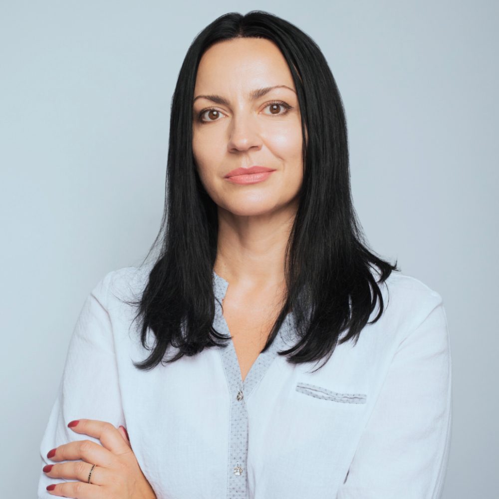 Portrait of a confident woman with long black hair, wearing a white blouse with a light gray trim, standing against a neutral gray background.