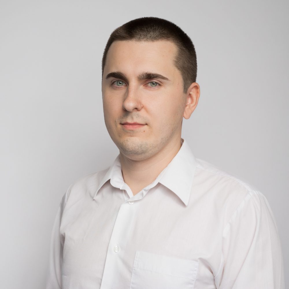 Young man with short dark hair, wearing a crisp white dress shirt, standing against a light gray background with a calm, neutral expression.